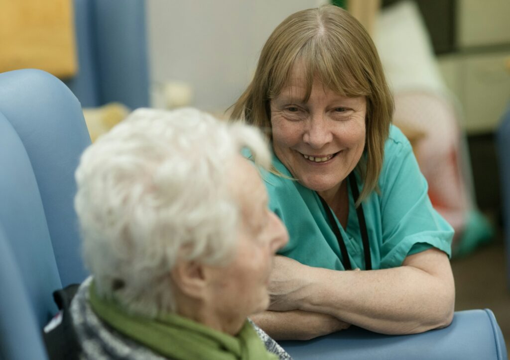 Smiling woman in green scrubs leaning towards an elderly person seated in a chair